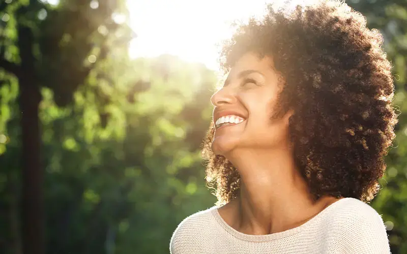 Mujer sonriente con cabello rizado mirando hacia arriba al aire libre, con luz solar brillante y fondo de árboles desenfocado.