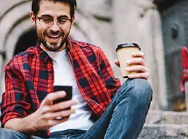Hombre joven sentado al aire libre, sonriendo mientras mira su celular y sostiene un vaso de café.