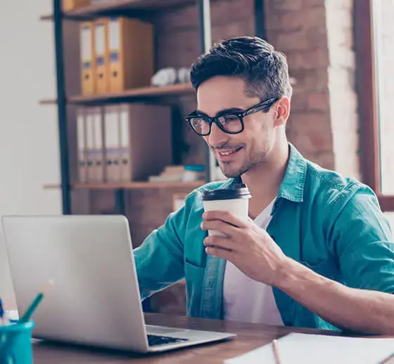 Hombre joven con gafas trabajando en su computadora portátil mientras sostiene un vaso de café en una mano.