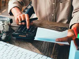 Persona usando una calculadora mientras revisa documentos financieros sobre un escritorio con teclado y frasco de monedas.