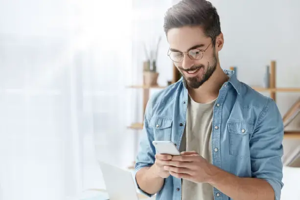Hombre joven con gafas y barba usando su celular, sonriendo mientras revisa algo en su dispositivo móvil, en un ambiente de trabajo con luz natural.