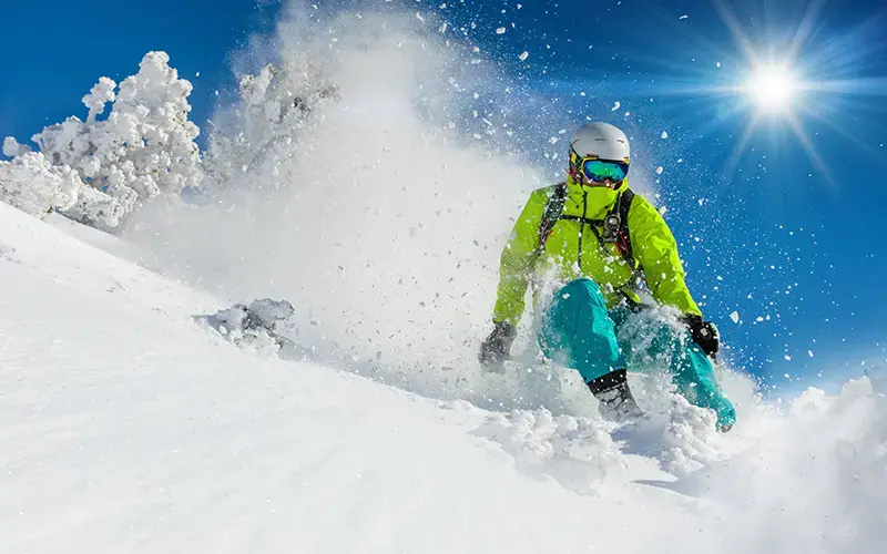 Snowboarder descendiendo por una pendiente nevada con equipo brillante bajo un cielo azul y soleado.