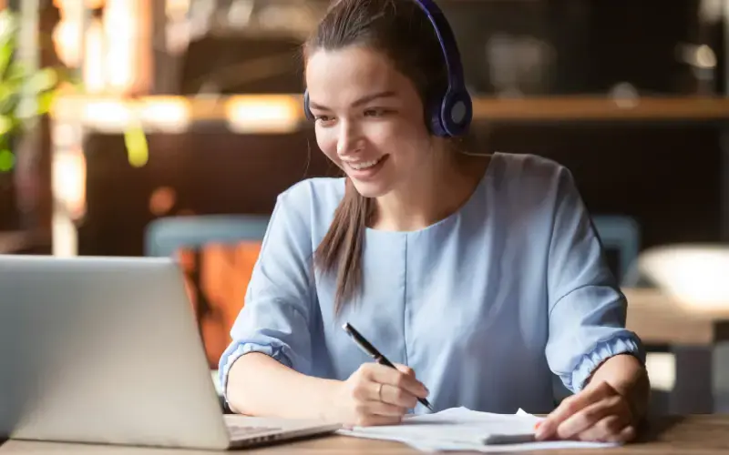 Mujer con audífonos escribiendo en una libreta mientras mira una computadora portátil.