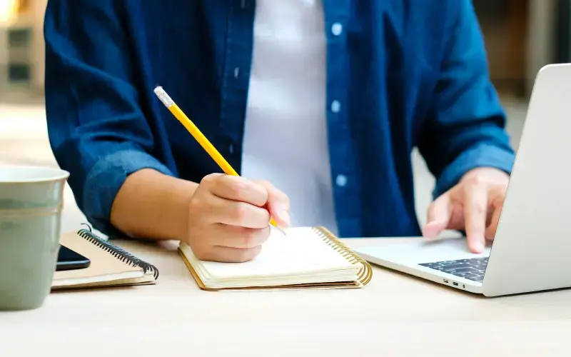 Persona escribiendo en una libreta con lápiz amarillo frente a una laptop, en un entorno iluminado.