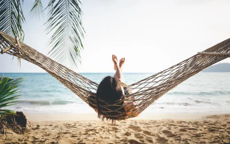 Mujer recostada en una hamaca en la playa, de espaldas, contemplando el mar bajo la sombra de una palmera.