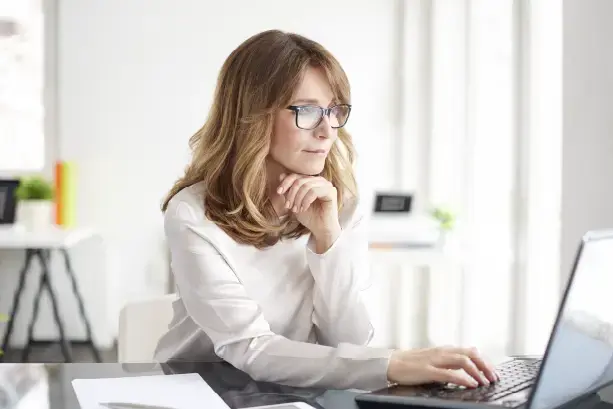 Mujer de mediana edad con gafas trabajando en una computadora portátil, concentrada en una oficina luminosa y moderna.