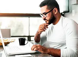 Hombre con barba y gafas trabajando concentrado en una laptop con taza de café al lado.