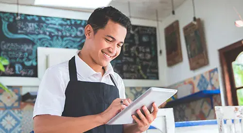 Joven emprendedor con delantal usando una tablet en el interior de su negocio decorado con pizarras y cerámica artesanal.