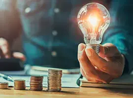 Persona sosteniendo un foco encendido junto a pilas de monedas apiladas sobre una mesa, simbolizando ideas financieras o ahorro energético.
