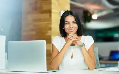 Mujer joven sonriente sentada frente a una computadora portátil en una oficina moderna con iluminación suave.