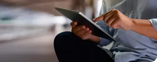 Persona usando una tableta digital con una mano mientras sostiene el dispositivo con la otra, en un entorno desenfocado.