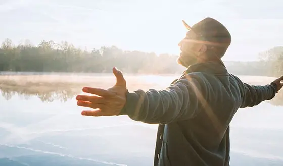 Persona con los brazos extendidos disfrutando del amanecer junto a un lago tranquilo.