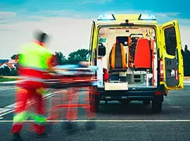 Paramédico empujando una camilla hacia una ambulancia con las puertas traseras abiertas y luces encendidas.