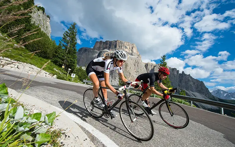 Dos ciclistas descendiendo por una carretera de montaña en un entorno natural con cielo azul y montañas al fondo.
