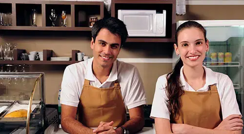 Dos emprendedores jóvenes atendiendo con amabilidad en su negocio propio, detrás del mostrador de una cafetería o tienda local.
