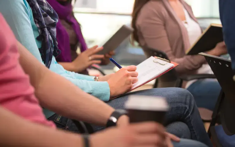 Grupo de personas sentadas tomando notas en una sala, algunas sosteniendo carpetas o tablets.