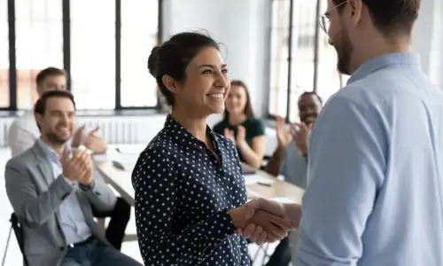 Mujer sonriente estrechando la mano de un compañero mientras colegas aplauden en una oficina
