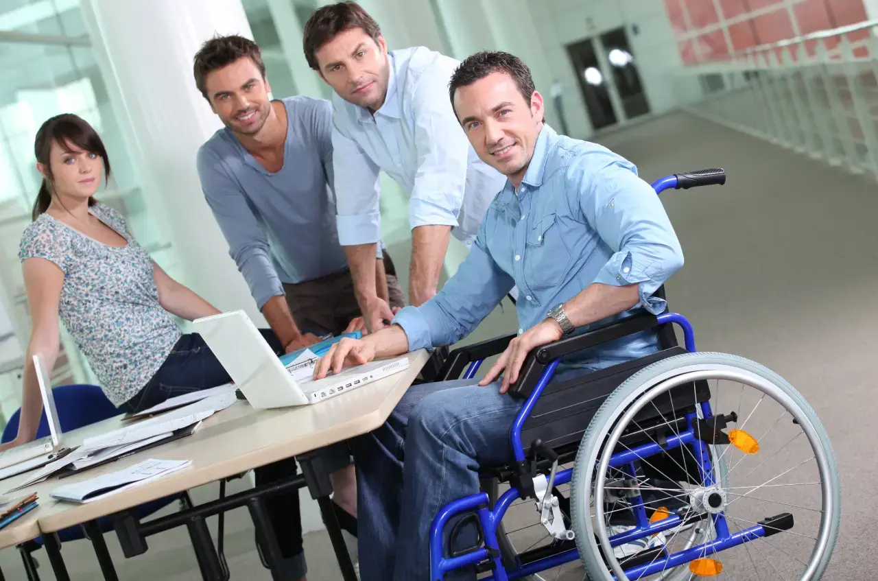 Hombre en silla de ruedas trabajando con su equipo en una oficina, todos reunidos alrededor de una mesa con computadoras portátiles