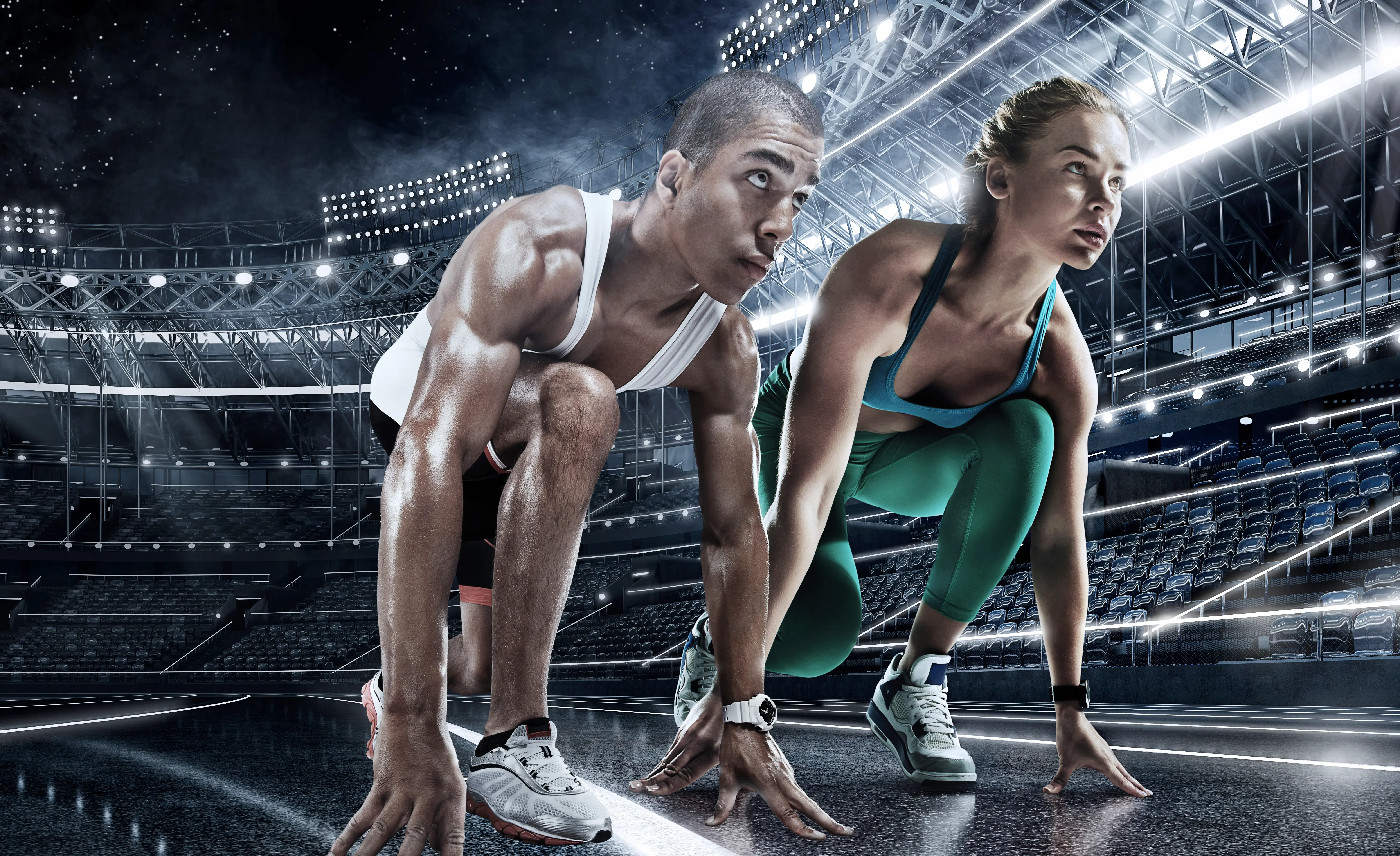 Hombre y mujer en posición de salida sobre una pista de atletismo dentro de un estadio iluminado, listos para correr.