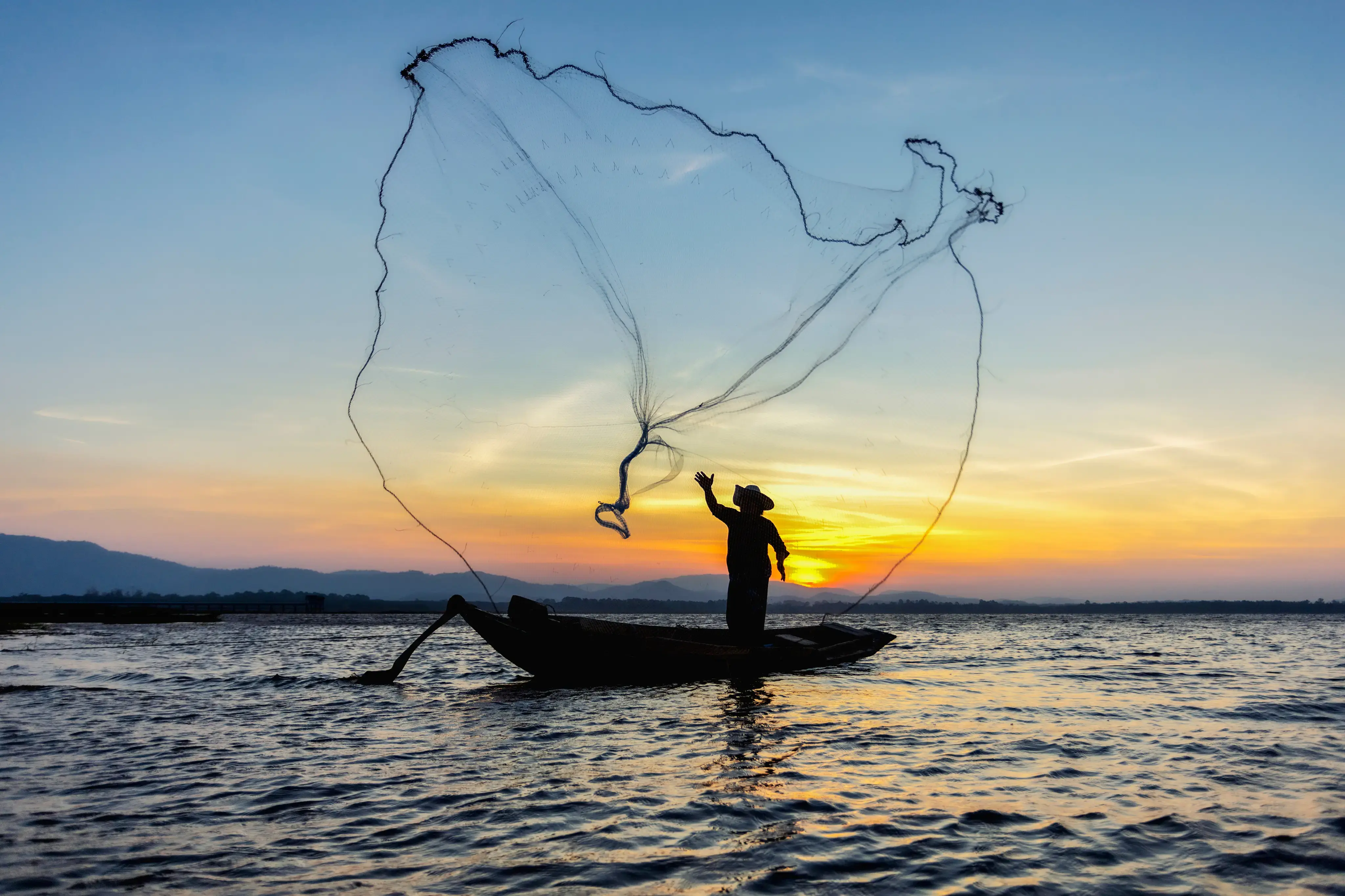 Pescador sobre una barca lanzando una red al atardecer en aguas tranquilas.