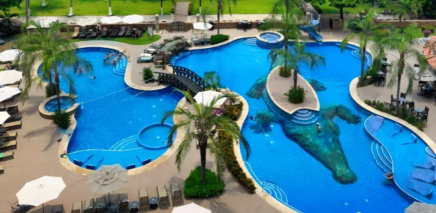 Vista aérea de piscina con forma irregular rodeada de palmeras, puentes y áreas de descanso