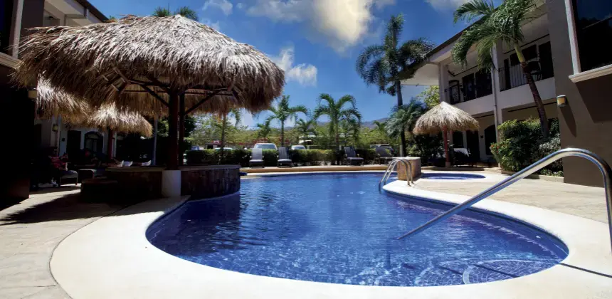 Piscina con sombrillas de palma y tumbonas en hotel rodeado de palmeras bajo cielo despejado