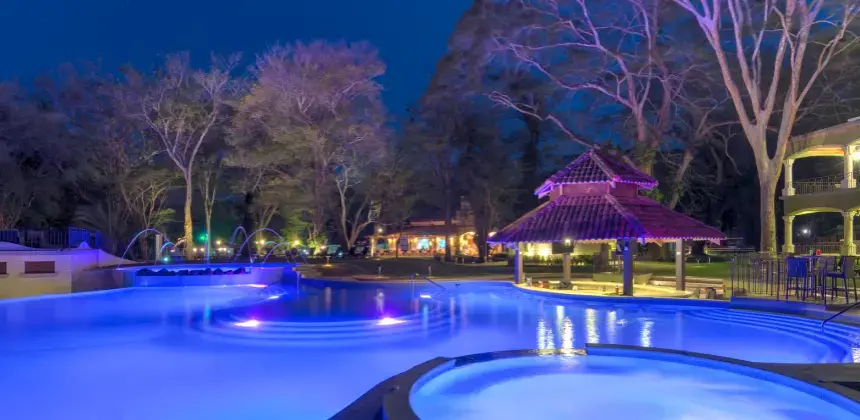 Piscina iluminada de noche con fuentes y un rancho tropical rodeado de árboles