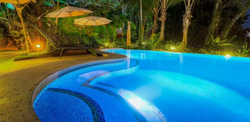 Piscina iluminada de noche rodeada de palmeras y sillas con sombrillas en un ambiente tropical