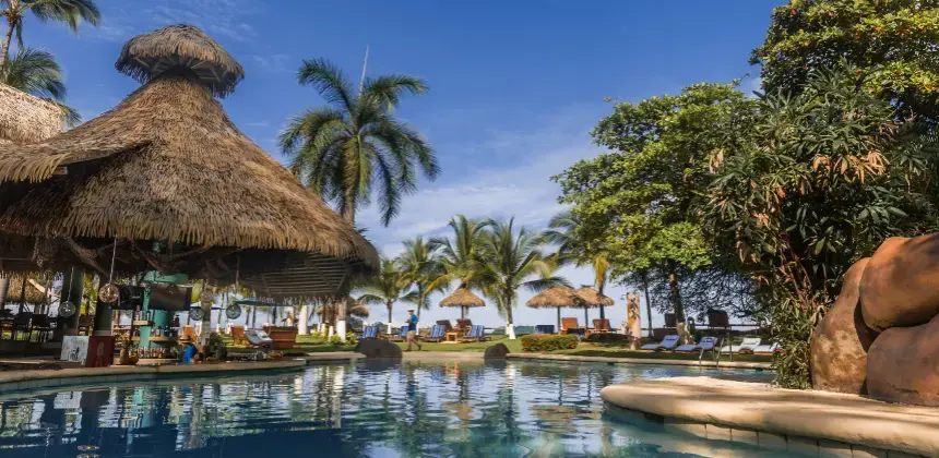 Piscina junto a la playa rodeada de palmeras y ranchos con techo de palma en un resort tropical