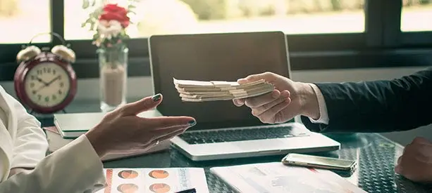 Mano entregando un fajo de billetes a otra persona en un entorno de oficina.