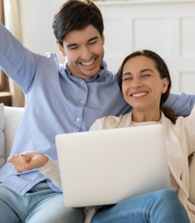 Una pareja feliz celebra frente a una computadora portátil, posiblemente por haber logrado una meta financiera o completado un trámite exitosamente.