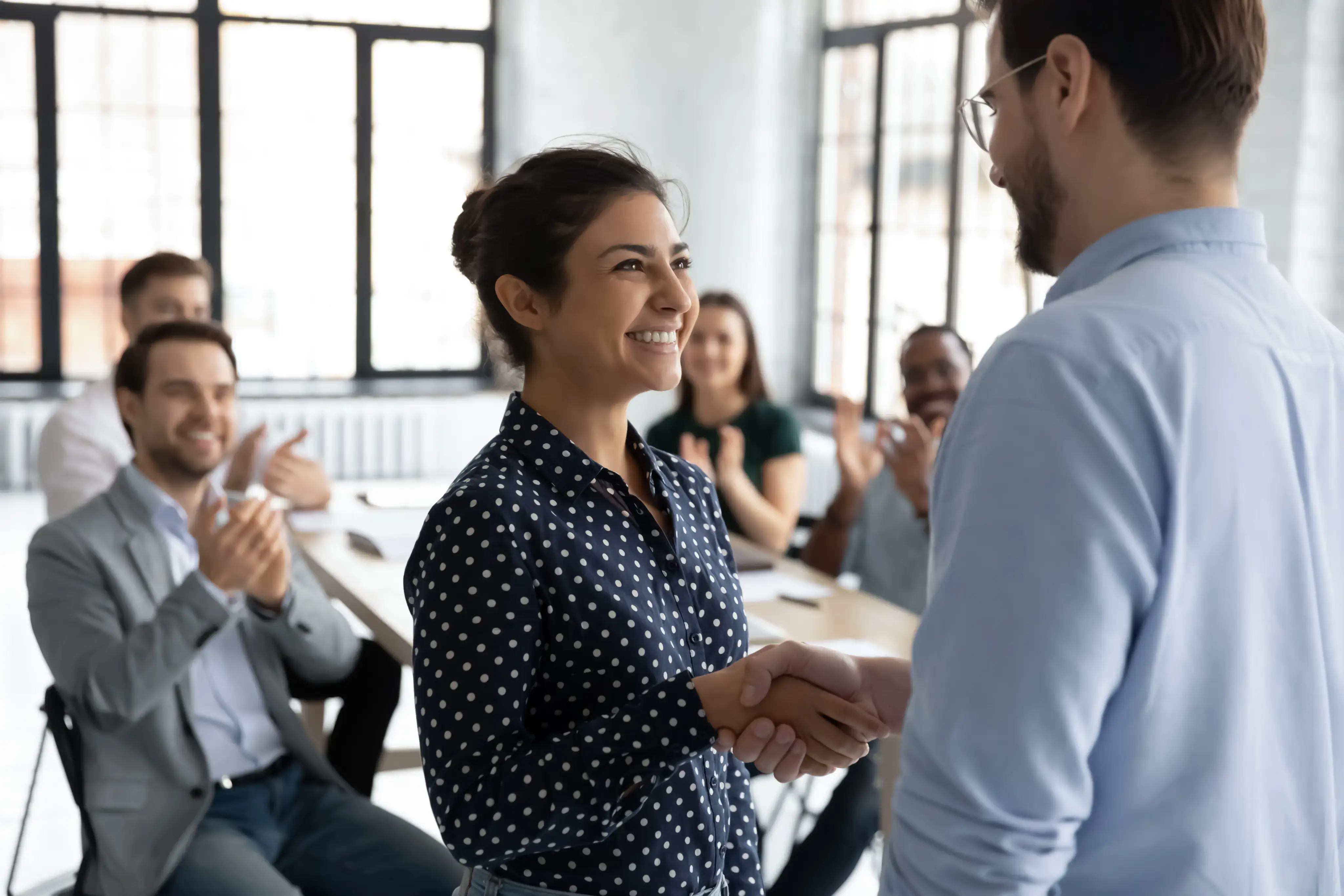 Mujer sonriente estrechando la mano de un colega mientras recibe reconocimiento en una reunión de trabajo.