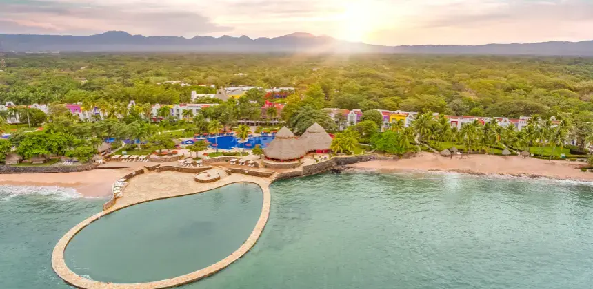 Vista aérea de un resort frente al mar con piscina natural y playa al atardecer.