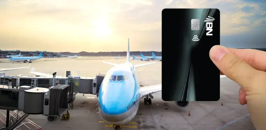 Persona sosteniendo una tarjeta BN negra frente a un avión en pista de aeropuerto al atardecer