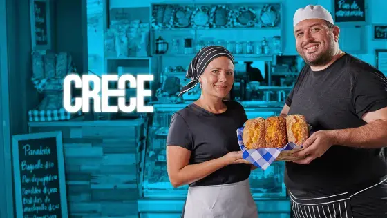 Hombre y mujer panaderos sonriendo mientras sostienen una bandeja con panes recién horneados, frente al mostrador de una panadería con la palabra 
