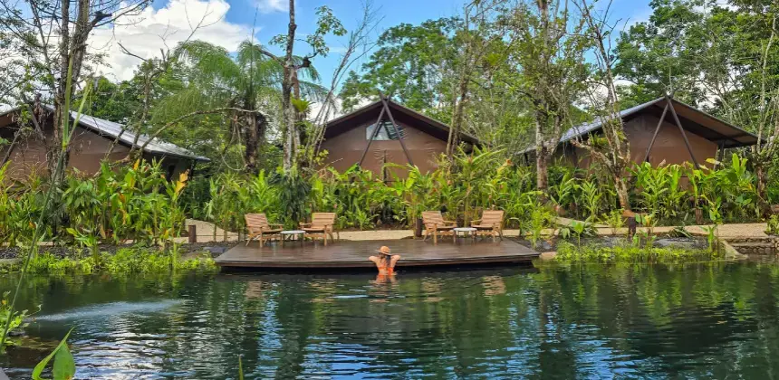 Persona disfrutando de una piscina natural rodeada de vegetación tropical y cabañas en Tocu Tent Camp