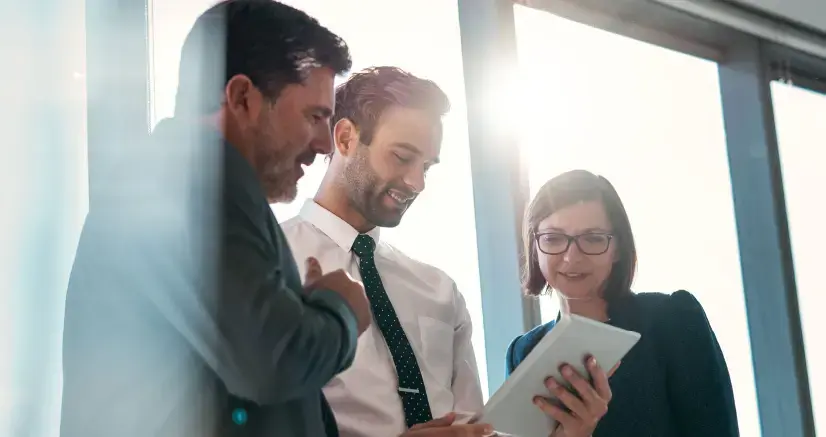 Tres personas de negocios observando una tableta digital mientras conversan en una oficina iluminada por el sol.