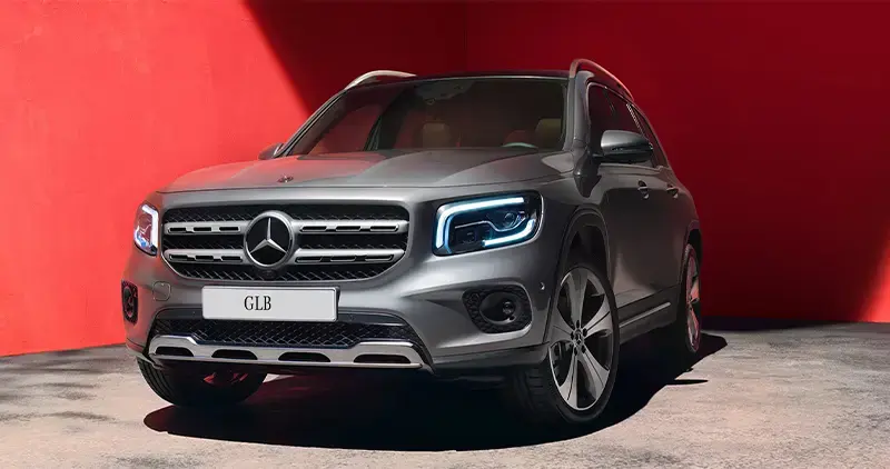Vehículo Mercedes-Benz GLB gris estacionado frente a una pared roja iluminada por luz solar.