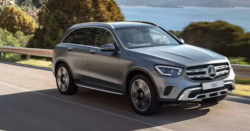 Vehículo Mercedes-Benz GLC gris circulando por carretera junto al mar con árboles y montañas al fondo.
