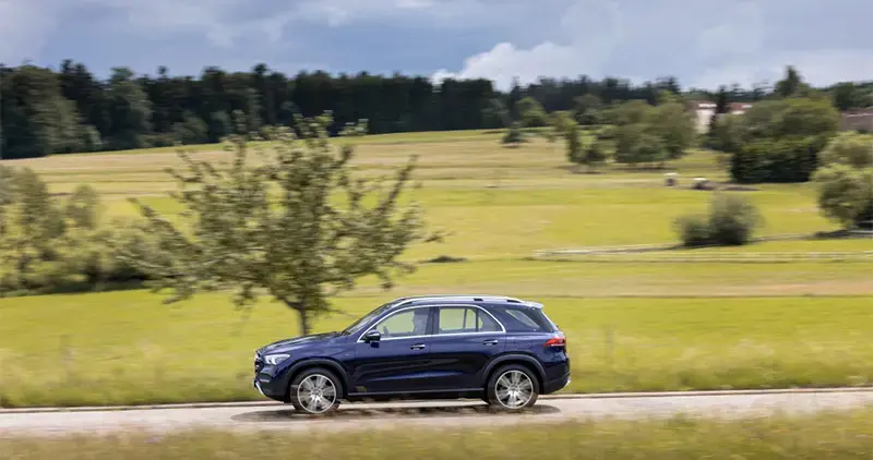 Vehículo Mercedes-Benz GLE 300 azul circulando por carretera rural entre campos verdes y árboles.