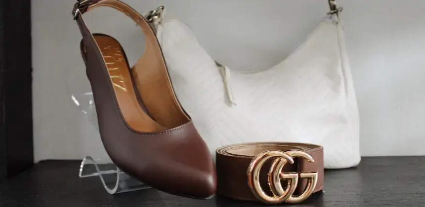 Zapatos marrones de tacón bajo junto a un cinturón con hebilla dorada y un bolso blanco sobre superficie oscura